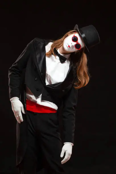 Portrait of male mime artist performing, isolated on black background. Man is standing straight tilt his head with his hand on the stomach.