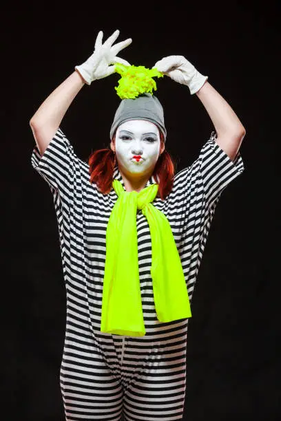 Portrait of female mime artist, isolated on black background. Young woman in striped clothes touching her funny bright hat.