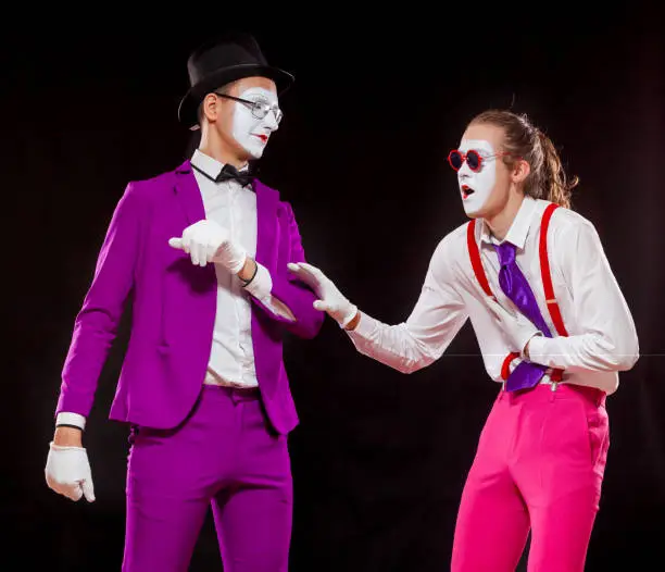 Portrait of two male mime artists, isolated on black background. A man in suspenders clutches his stomach and reach for a man in purple suit.