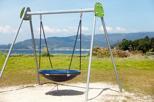 Empty swing, child's playground toy, shadow and beach sand,  ria de Vigo landscape in the background, Rias Baixas, Pontevedra province, Galicia, Spain.