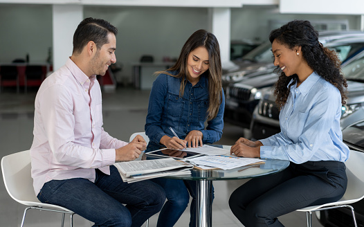 Happy Latin American couple buying a car from a saleswoman at a car dealership and signing a contract