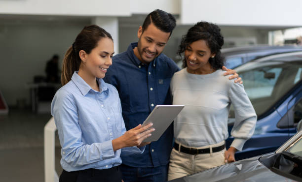 Saleswoman at a car dealership talking to a happy couple Portrait of a saleswoman at a car dealership talking to a happy couple and showing them info on a tablet computer - business concepts car dealership stock pictures, royalty-free photos & images