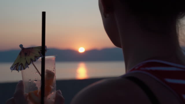 Girl drinking fresh mojito with drinking straw, looking in distance and enjoying amazing marine sunset. Silhouette of young teenager at the sea beach relaxing and drinking beverage. Summer vacation memories, nostalgia concept