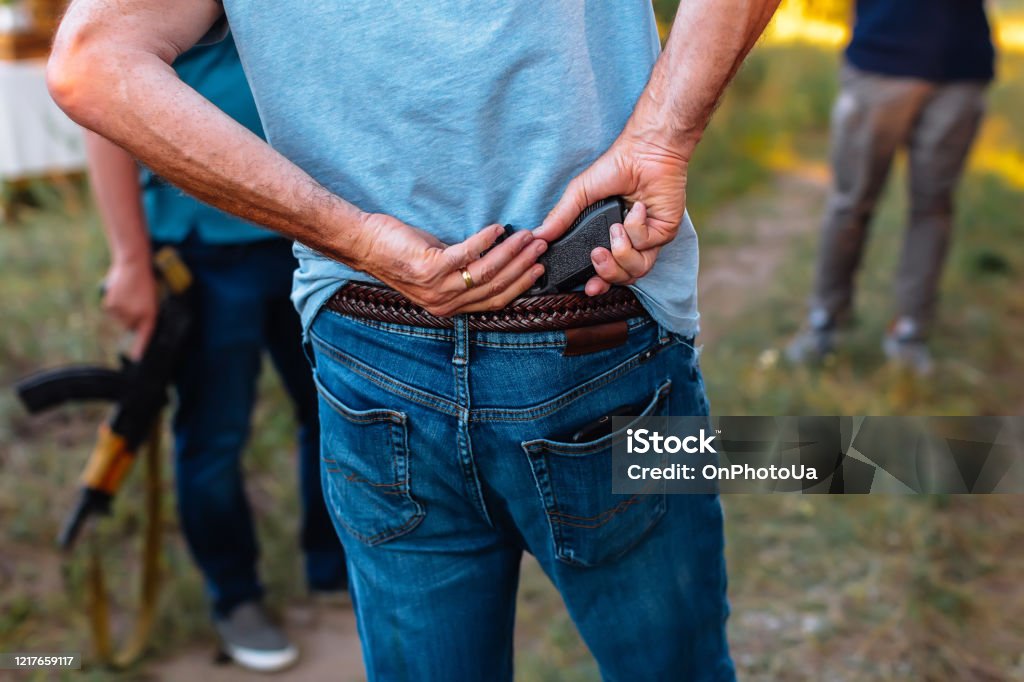 man hides a pistol behind his back under the belt of jeans man hides a pistol behind his back under the belt of jeans with group of criminals Gun Stock Photo