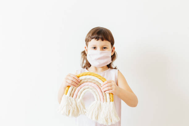 let's all be well. child at home wear reusable textile mask hold textile handmade rainbow. stock photo