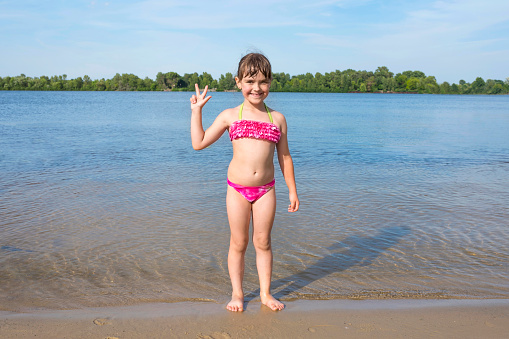 A little happy girl in a pink bathing suit standing on the bank of a river on a sunny summer day.  Beach season.