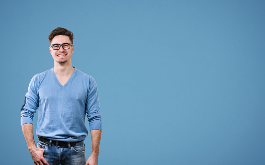 Young handsome man in sweater and eyeglasses looking at camera standing on blue background.