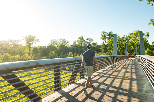 This is a photograph of a man walking across a pedestrian bridge in Buffalo Bayou park in downtown Houston, Texas on a sunny spring day.