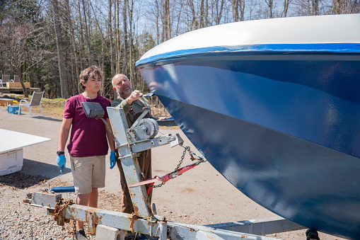 A father and his teen-aged son painting an old boat they are restoring in the driveway of their home.