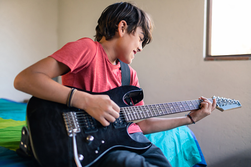 Latinx pre-adolescent child cheerfully playing electric guitar in his room
