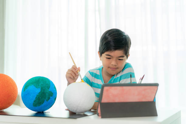 istock Asian elementary schoolboy painting the moon in science class learning about the solar system via video conference with teacher and other classmates at home, Homeschooling and distance learning. 1217687563