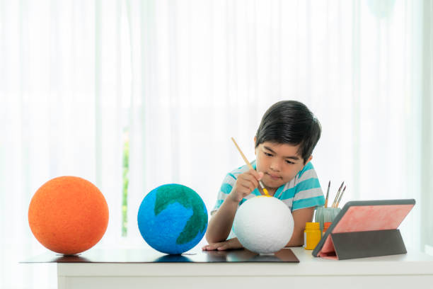 istock Asian elementary schoolboy painting the moon in science class learning about the solar system via video conference with teacher and other classmates at home, Homeschooling and distance learning. 1217687565