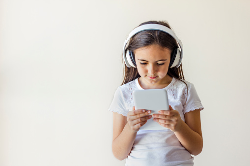 Beautiful little girl holding a tablet in her hands. The girl is wearing headphones. Online Learning.  Portrait isolated on a grey background.