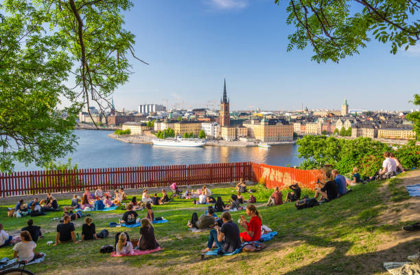 Young people spend time (sit, drink, eat) on lawn with green grass and trees around on Sodermalm island with view of old historical town Riddarholmen and Gamla Stan stock photo