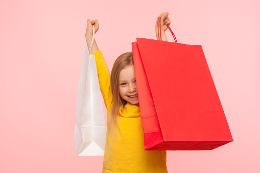 Portrait of happy carefree little girl lifting up large packages and smiling happily, excited about shopping in children store, Black Friday sales. indoor studio shot isolated on pink background