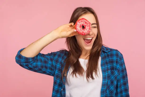 Photo of Junk food and confectionery. Portrait of glad playful girl in checkered shirt looking through doughnut