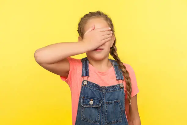 Photo of Portrait of scared upset little girl with braid in denim overalls covering eyes with hand, child feeling shame