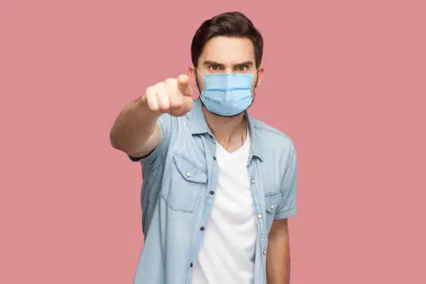Hey you. Portrait of serious man with surgical medical mask in blue casual style shirt standing pointing and looking at camera with angry face. indoor studio shot, isolated on pink background.