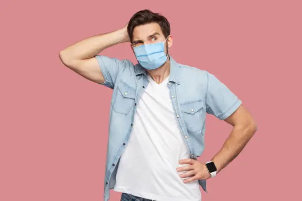 Portrait of young man with surgical medical mask in blue casual style shirt standing, scratching his head, looking away and thinking what to do. indoor studio shot, isolated on pink background.