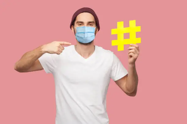 Photo of Portrait of happy young hipster man with surgical medical mask in white shirt and casual hat standing, holding yellow hashtag and pointing finger to it.