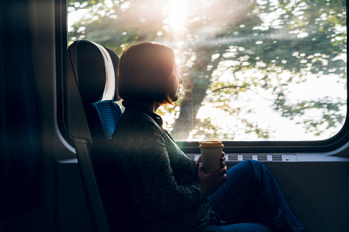 A middle aged woman sitting by the window of a commuter train with a laptop and coffee in a reusable bamboo cup in hand. The concept of remote work, business trips, freelancer, Tourist solo travel