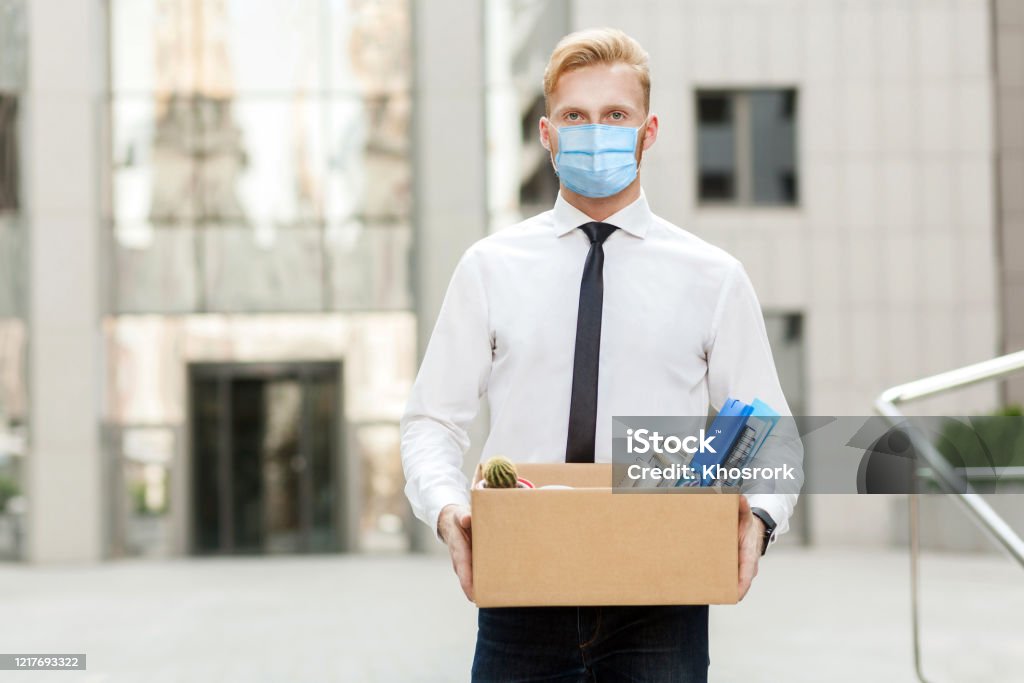 lose job, Youre fired. Unhappy business man with surgical medical mask going out with cardboard, looking at camera and feeling looser. lose job, Youre fired. Unhappy business man with surgical medical mask going out with cardboard, looking at camera and feeling looser. Outdoor shot. business, working and health care concept. Downsizing - Unemployment Stock Photo