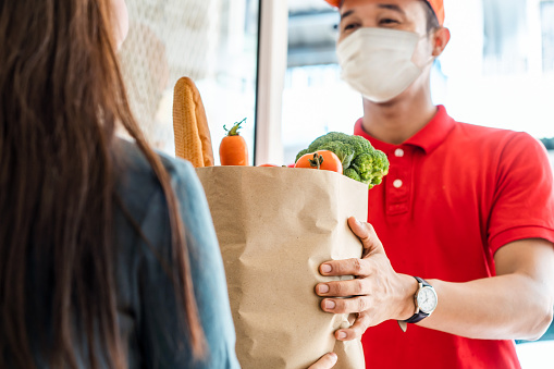 Asiático entregar hombre usando máscara facial en rojo uniforme manipulación bolsa de comida, fruta, verduras dar a la mujer cliente en frente de la casa. Servicio de entrega de correos y comida exprés durante covid19. photo