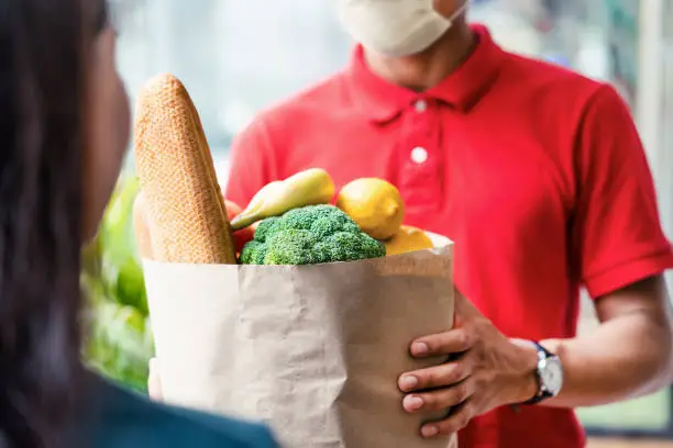 Asian deliver man wearing face mask in red uniform handling bag of food, fruit, vegetable give to female costumer in front of the house. Postman and express grocery delivery service during covid19.