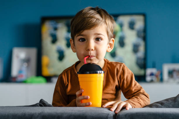 porträt von kleinen niedlichen kaukasischen jungen drei oder vier jahre alt sitzen auf dem bett oder sofa zu hause hält eine plastiktasse mit stroh trinken saft soda allein am tag suchen kamera vor dem fernseher - 3 4 years cute beauty beautiful stock-fotos und bilder