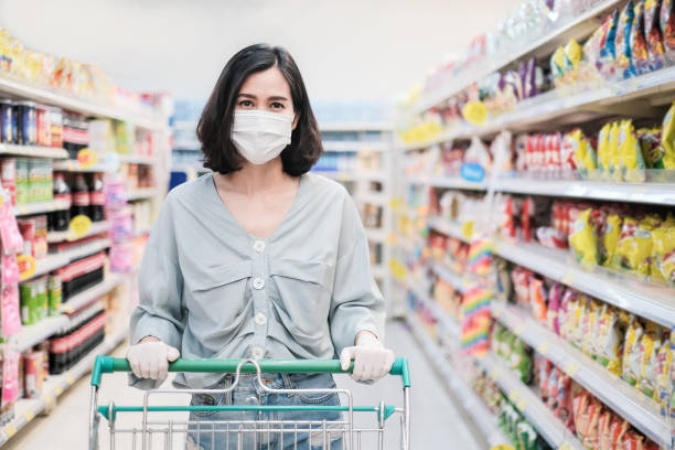 asian woman wearing face mask and rubber glove push shopping cart in suppermarket departmentstore. girl choosing, looking grocery things to buy at shelf during coronavirus crisis or covid19 outbreak. - asian ethnicity shopping mall supermarket store imagens e fotografias de stock