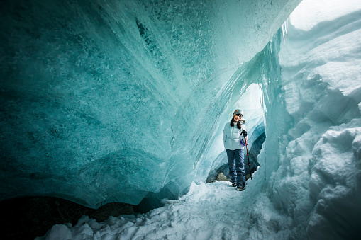 Female explorer wearing ski gear and helmet touching ice in glacial cave.