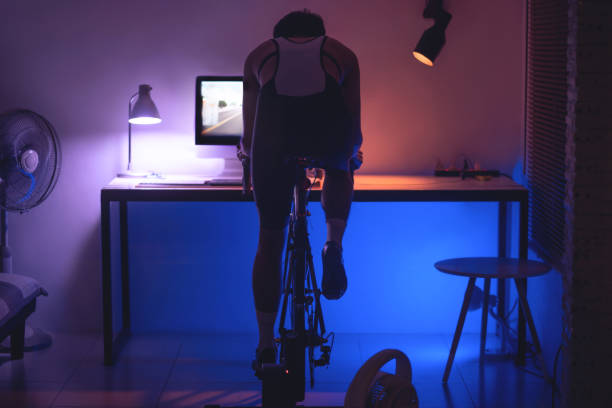 homem asiático pedalando no treinador de máquinas que ele está exercitando em casa à noite.ele joga jogo de bicicleta online - short game - fotografias e filmes do acervo