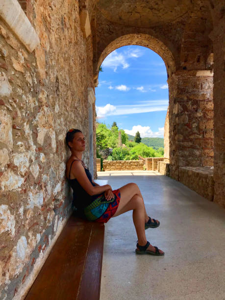 Female tourist sitting on a bench under historic arcades with a window like idyllic view to landscape and blue sky on a summer day Woman taking a rest from visiting the Pantanassa Monastery (Greek: Μονή Παντανάσσης) Mystras or Mistras (Greek: Μυστρᾶς/Μιστρᾶς), Peloponnese or Peloponnisos, Greece older women short skirts stock pictures, royalty-free photos & images