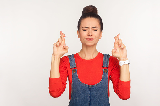 Portrait of imploring girl with hair bun in overalls holding fingers crossed for good luck, dreaming of fortune, waiting miracle, making wish with expression of great desire. studio shot, isolated