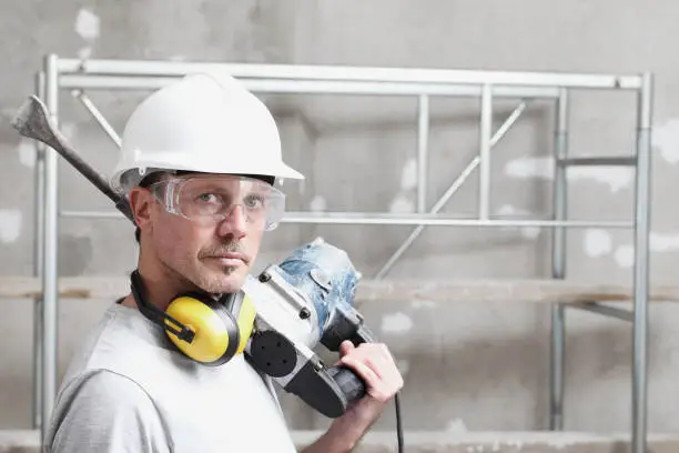 portrait of man construction worker with jackhammer with safety hard hat, hearing protection headphones and protective glasses. look at the camera isolated on interior building site background