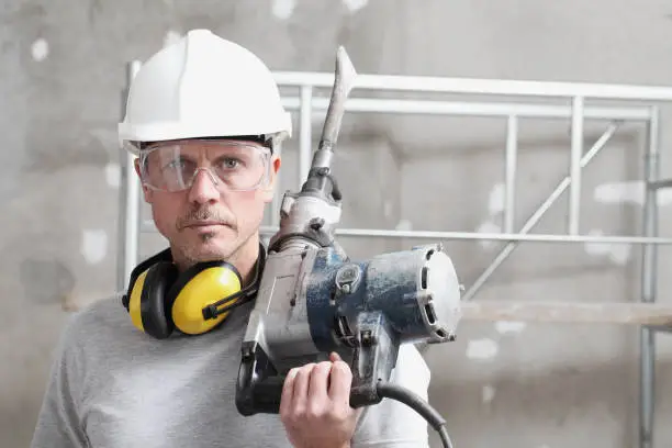 portrait of man construction worker with jackhammer with safety hard hat, hearing protection headphones and protective glasses. look at the camera isolated on interior building site background