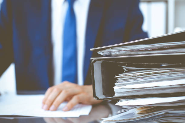 istock Binders with papers waiting to be processed with businessman or bookkeeper back in blur. Accounting planning budget. Audit, insurance and business concept 1217736214