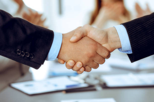 istock Business people shaking hands at meeting while theirs colleagues clapping and applauding. Group of unknown businessmen and women in modern white office. Success teamwork, partnership and handshake concept 1217736215