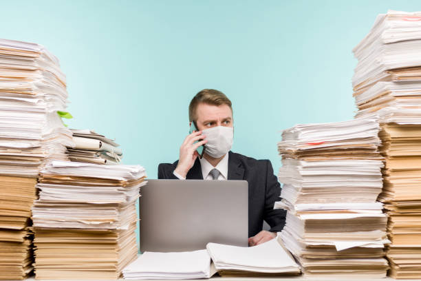A male accountant or company manager works in an office in a pandemic in view of the accumulated paper work. A protective medical mask is on the face. On the desktop are large stacks of documents. A male accountant or company manager works in an office in a pandemic in view of the accumulated paper work. A protective medical mask is on the face. On the desktop are large stacks of documents. - image bureaucracy photos stock pictures, royalty-free photos & images