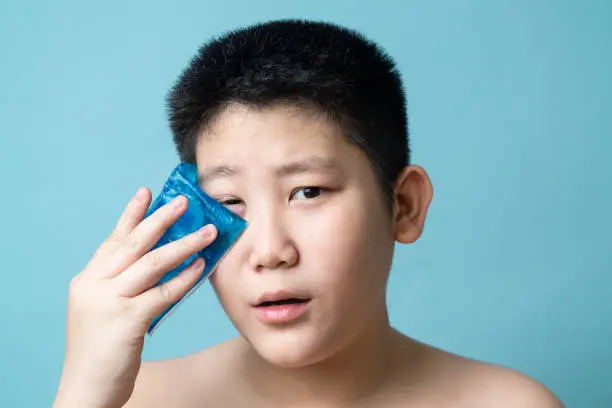 Asian preteen boy using cold compress gel on bruises face on blue background.