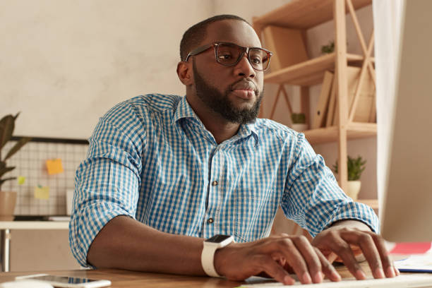 Busy day. Smart young afro american man in glasses working on the computer while sitting at his working place at home. Afro american businessman working remotely at home