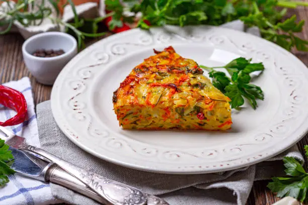 Piece of vegetable Kugel from zucchini, carrots, potatoes, garlic and turmeric in a white plate, selective focus. Dish of traditional Jewish cuisine.