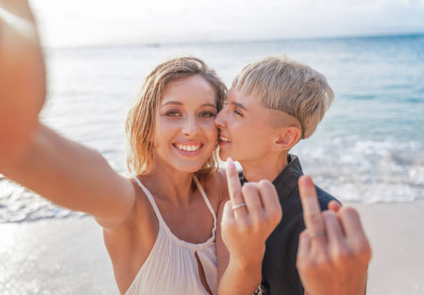 duas lindas jovens mulheres celebram um casamento lésbico no caminho durante a lua de mel, retrato de primavera de verão, tirar uma selfie em um smartphone e mostrar anéis nos dedos - honeymoon wedding beach couple - fotografias e filmes do acervo