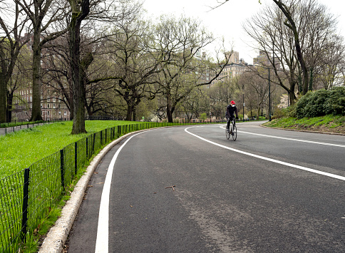 New York, NY, USA - April 8, 2020: A cyclist wears a red Yankees cap and a mask as he bikes on the East Drive of a very empty Central Park. The deserted park and his mask reflect the ongoing coronavirus pandemic. To the left through the trees can be seen the building lining Fifth Avenue on Manhattan's upper east side.