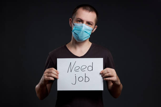 porträt eines mannes in einer medizinischen maske mit einem zeichen für notjob auf dunklem hintergrund, coronavirus-infektion - job search job sign cardboard stock-fotos und bilder