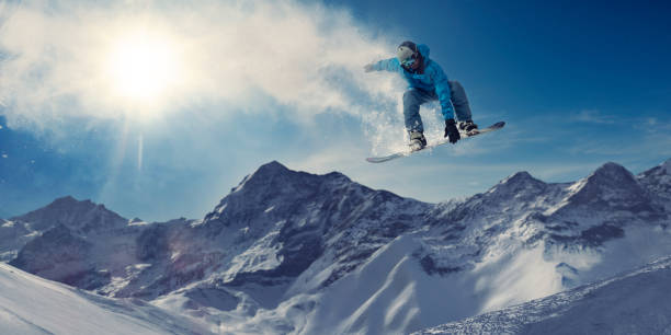 Extreme Snowboarder In Massive Big Air Jump In Snowy Mountains A male snowboarder in mid air during an extreme high jump in a generic location with snow covered mountains on bright day. The snowboarder is wearing blue toned snow-roof trousers and jacket, safety helmet and goggles. He leaves a snow vapour trail behind him, which the sun is visible through. snowboarding stock pictures, royalty-free photos & images