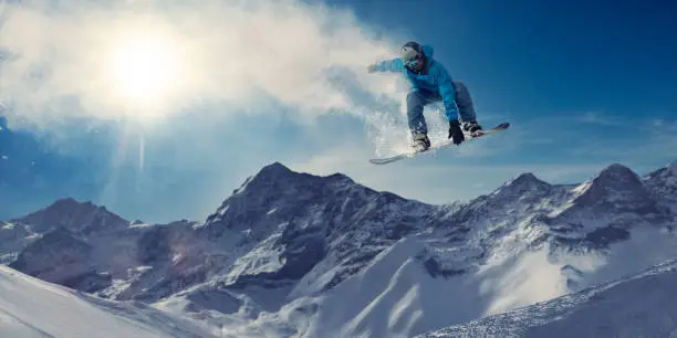 A male snowboarder in mid air during an extreme high jump in a generic location with snow covered mountains on bright day. The snowboarder is wearing blue toned snow-roof trousers and jacket, safety helmet and goggles. He leaves a snow vapour trail behind him, which the sun is visible through.