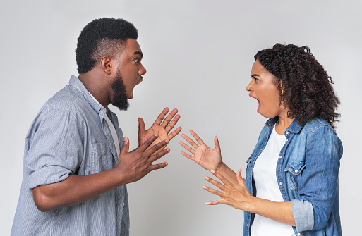 Couple Arguing. Angry Black Man And Woman Shouting At Each Other, Standing On Light Background In Studio, Side View