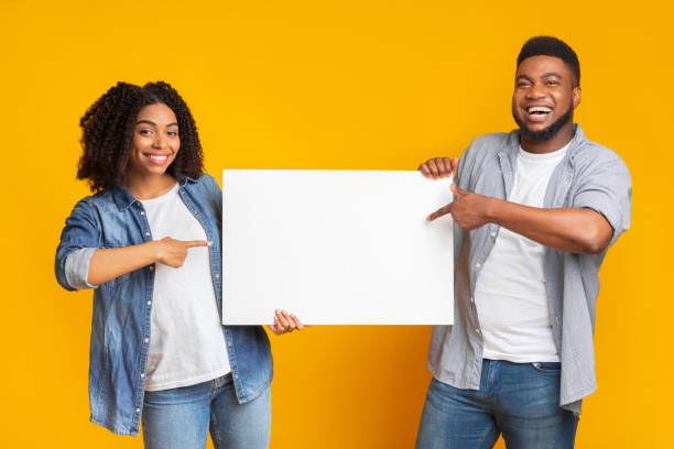 istock Cheerful black couple holding and pointing at blank advertisement placard 1217794152