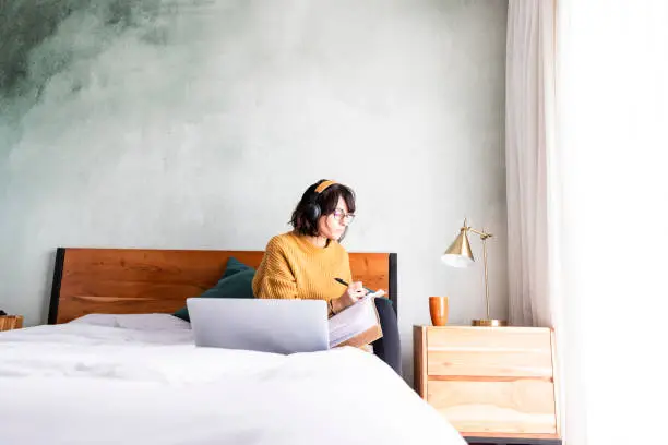 Photo of Wide shot woman taking notes while in video conference working in bedroom